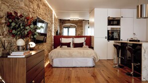 Ein Schlafzimmer mit Steinwand, einem hölzernen Schminktisch und einem Bett mit weißer Bettwäsche, sowie einer Küchenzeile mit Mikrowelle und Backofen.