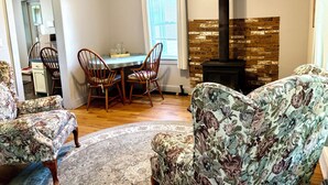 Een woonkamer met een open haard, fauteuils met een bloemmotief, een eettafel en een vloerkleed.