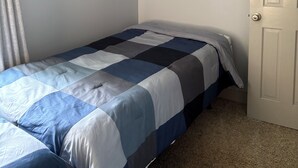 Een enkel bed met een blauw-witte quilt in een kamer met lichtgrijze wanden en een gesloten deur.