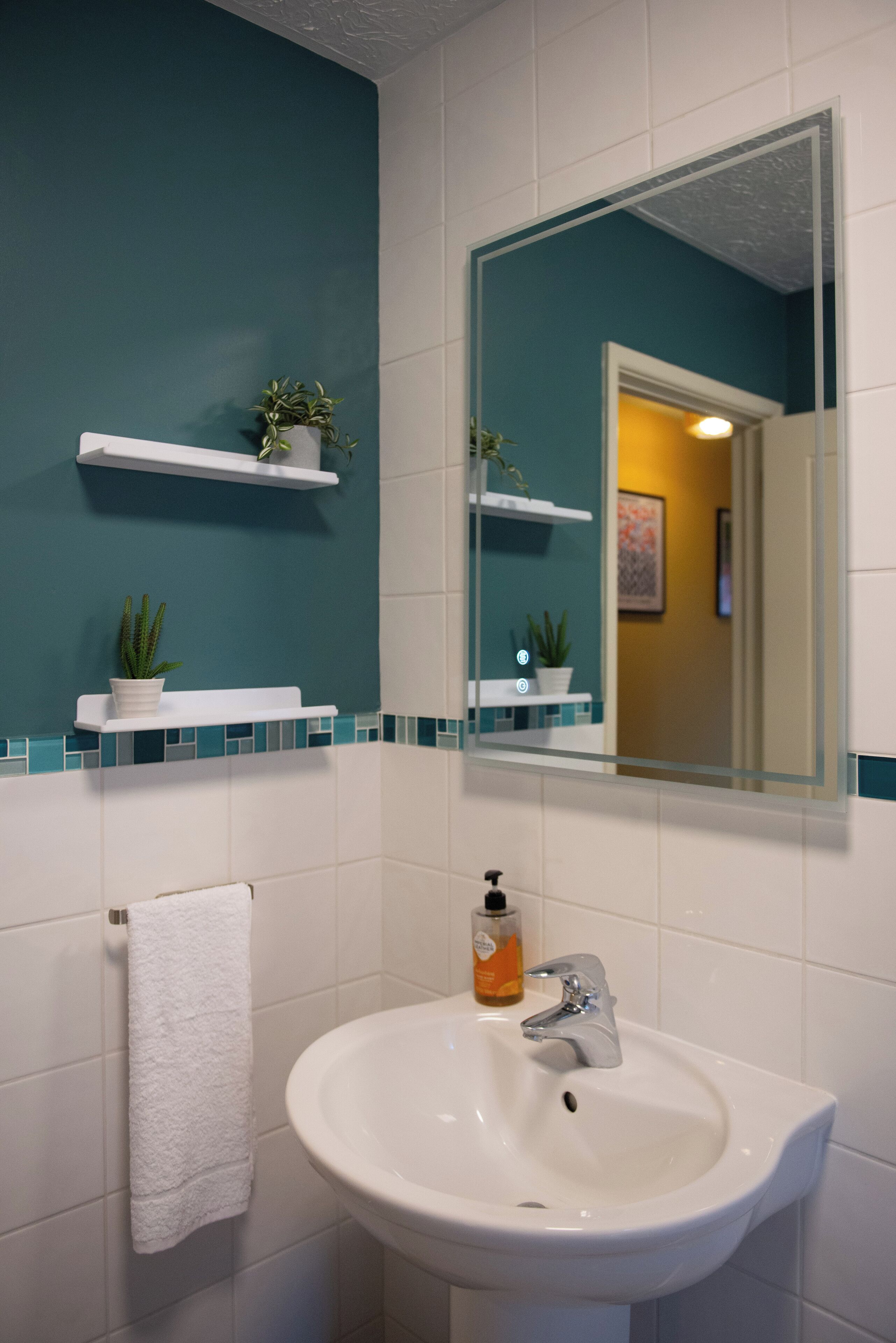 Een badkamer met een witte wastafel, wanden in turquoise en witte tegels. Er zijn twee witte planken met potplanten.
