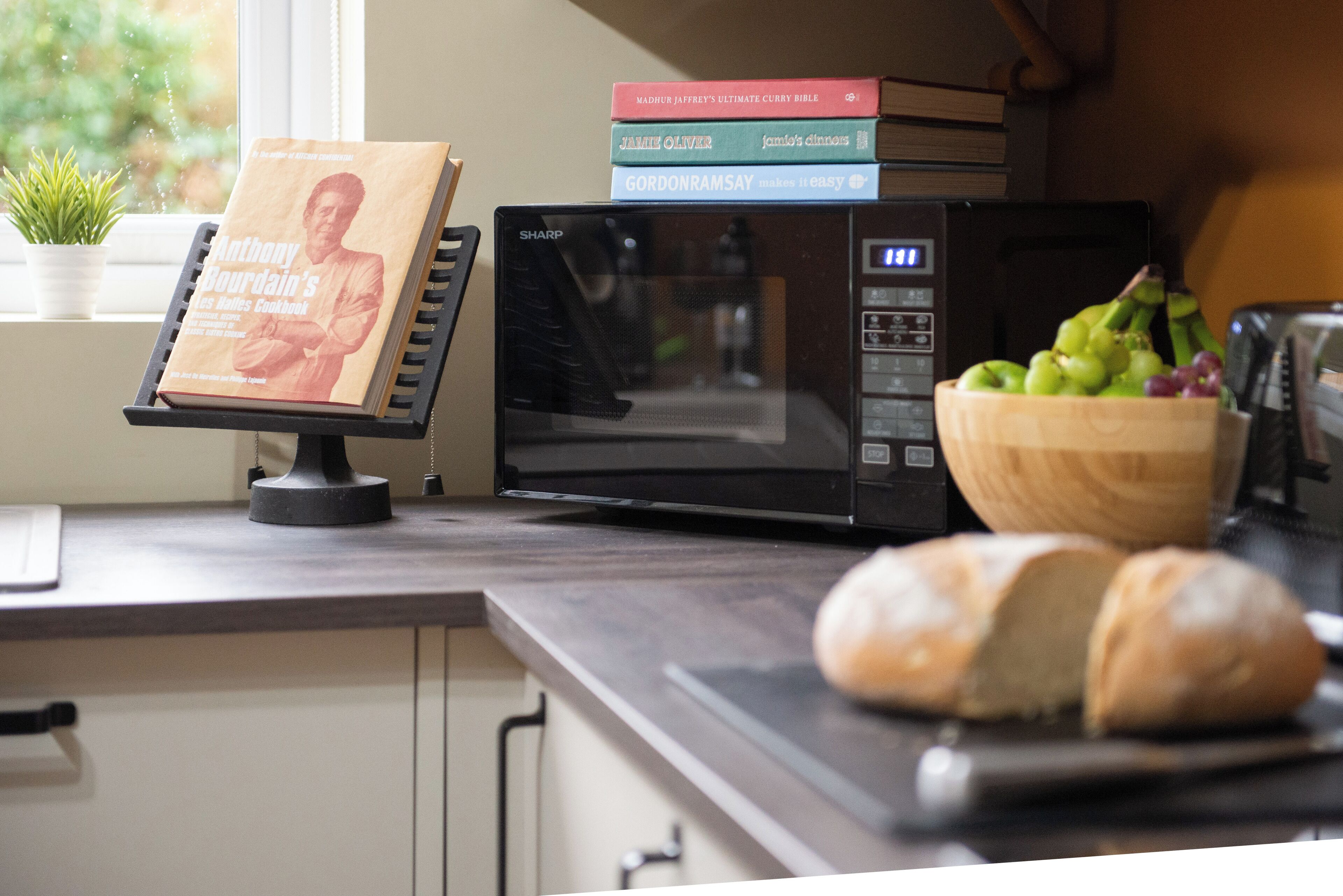 Een keukenblad met een magnetron, een boek getiteld 'Anthony Bourdain's Les Humbles Cookbook', een schaal druiven en een brood.