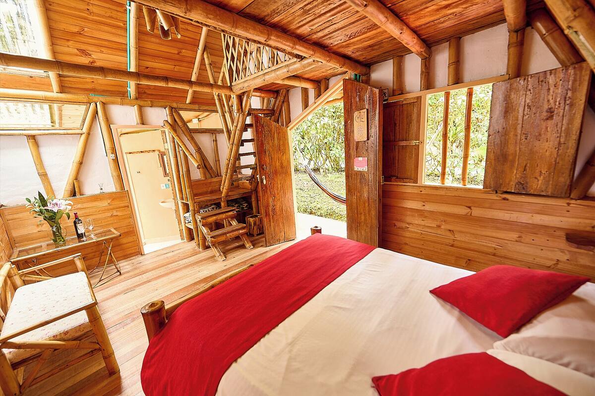 Une pièce de cabane en bois comprenant un lit, une chaise, une petite table avec une bouteille et un verre, et une fenêtre donnant sur un paysage verdoyant.
