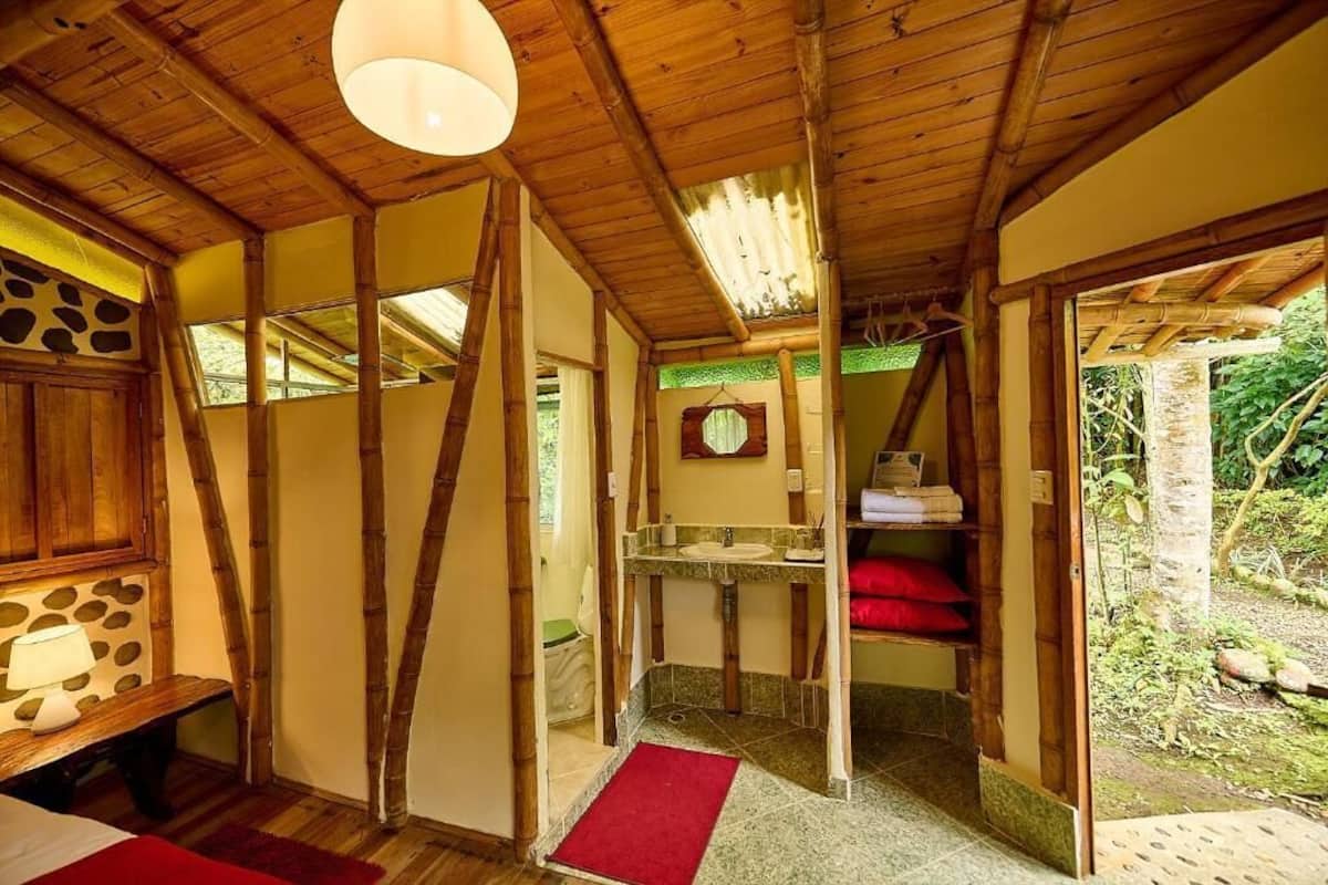Une pièce rustique en bois, avec un lit, un banc, une salle de bain dotée d’un lavabo et de serviettes rouges, et une vue sur la verdure à l’extérieur.