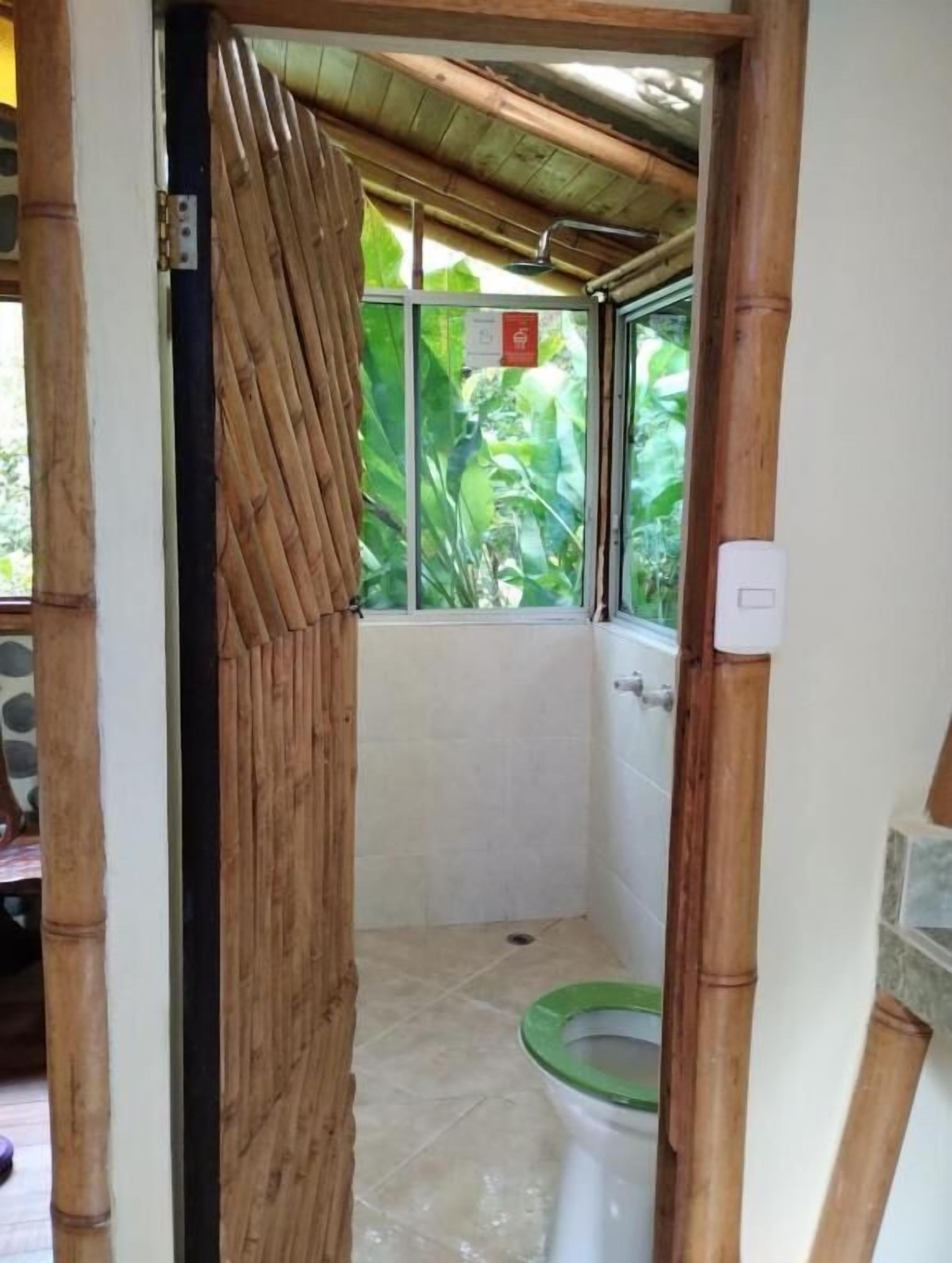 Un baño con puerta de bambú, inodoro con asiento verde y ventana con vista a follaje.