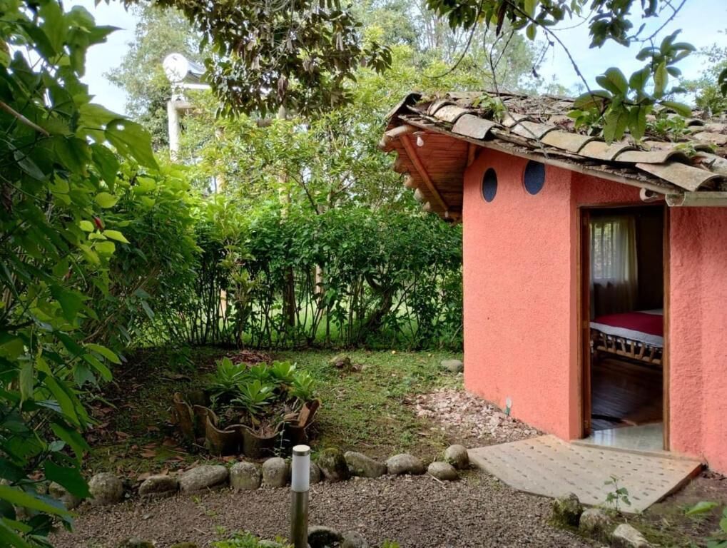 Una construcción pequeña con techo rosa, que tiene una cama en su interior, rodeada de exuberante vegetación y un sendero de grava.