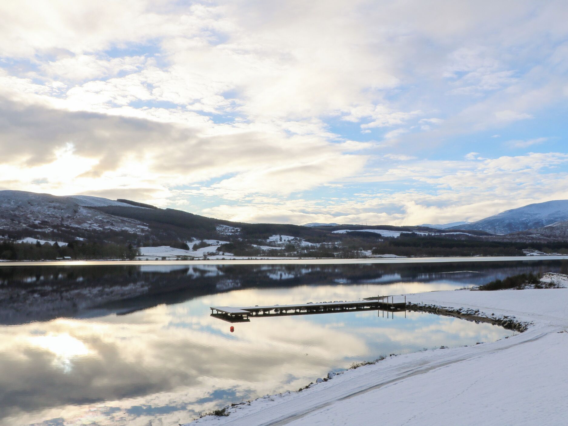 Ett snötäckt landskap med en sjö, en träbrygga och berg i bakgrunden.