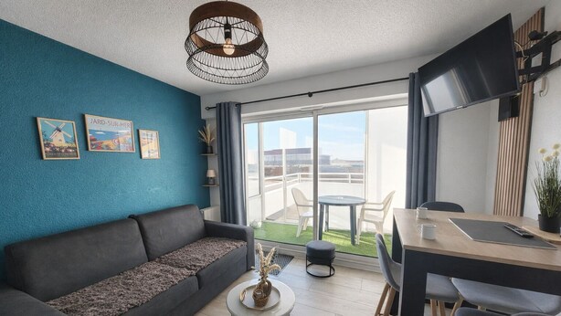 A modern living room with a dark sofa, a round coffee table, and a dining area with a table and chairs. There is a large window providing a view of the outdoors. - Cosy seaside studio in Jard-sur-Mer - Beach 50 m away, private parking (Jard-sur-Mer)