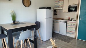 A compact kitchen with a white refrigerator, a small dining table with chairs, a potted plant, and a decorative vase on a side table. - Cosy seaside studio in Jard-sur-Mer - Beach 50 m away, private parking (Jard-sur-Mer)