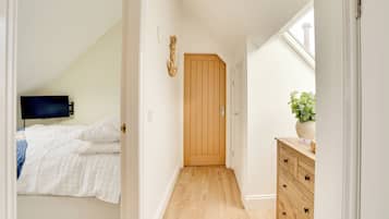 A narrow hallway with a wooden door, a bed with white bedding, a dresser with a plant, and a TV in the adjacent room.