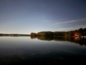 Ein ruhiger See im Abenddämmerung mit bewaldeter Uferlinie und einem kleinen Haus mit Lichtern.