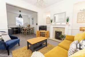 A living room with a sofa, a coffee table, a TV, a fireplace, and a dining area with chairs and a table. - Ivydene - 3 bedroom house -Tenby (Tenby)