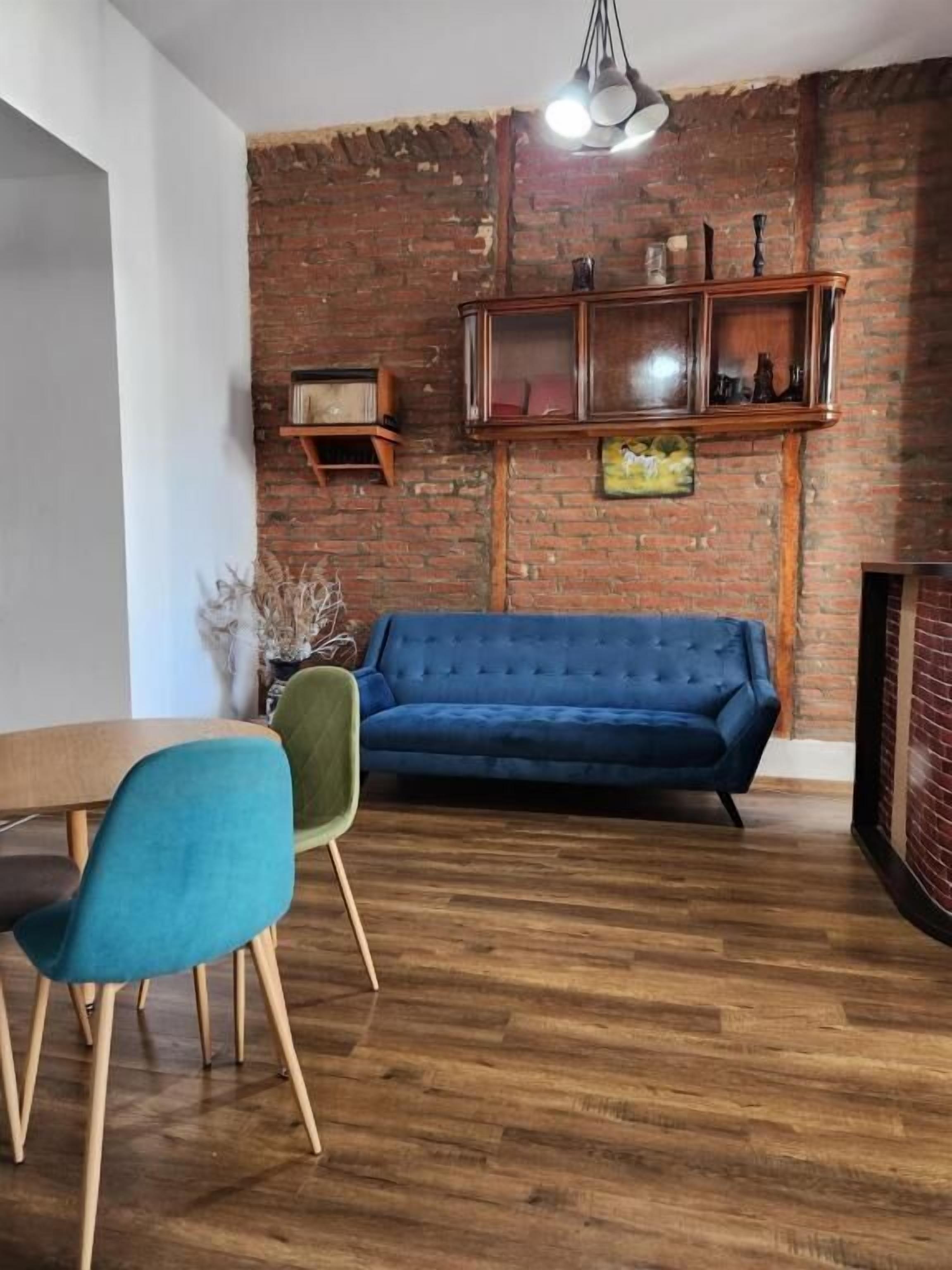 A room with a blue sofa, wooden dining table, and chairs, a brick wall, and a hanging light fixture.