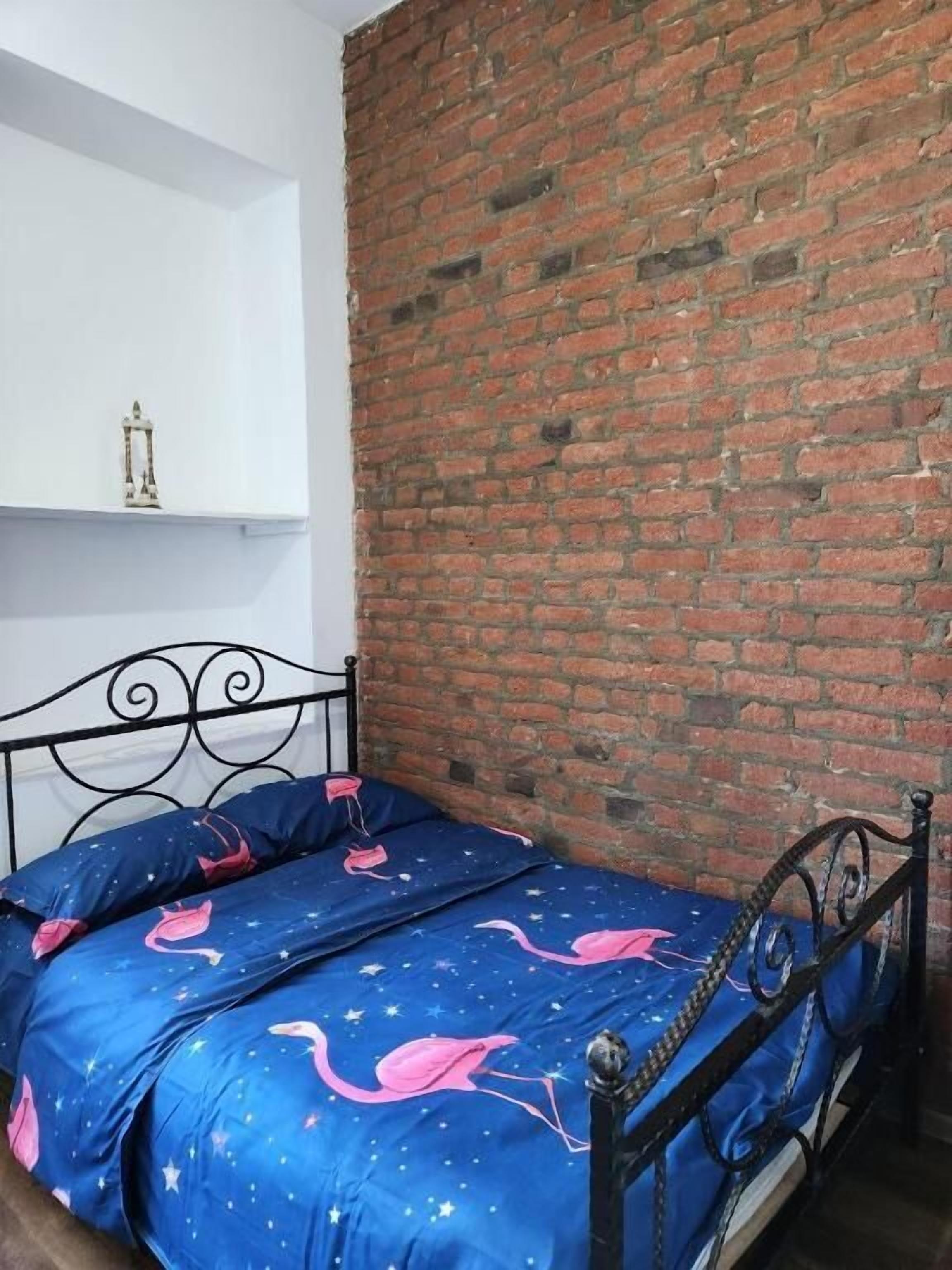 Quarto com parede de tijolos, estrutura de cama em metal e colcha azul estampada com flamingos rosa.