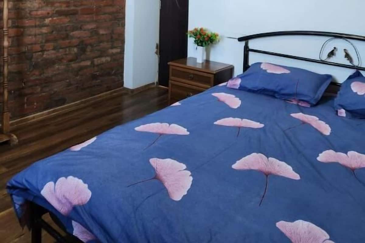 A bedroom with a brick wall, a bed with a floral bedspread, a nightstand with a vase of flowers, and a wooden floor.