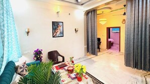 A modern living room with a sofa, a coffee table, and decorative plants. There is a painting on the wall and a chandelier in the background. - A Little Peaceful Retreat – Your Cozy Home Away From Home (Jaipur)