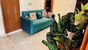 A teal sofa with patterned cushions, a wooden door, and large green plants. - A Little Peaceful Retreat – Your Cozy Home Away From Home (Jaipur)