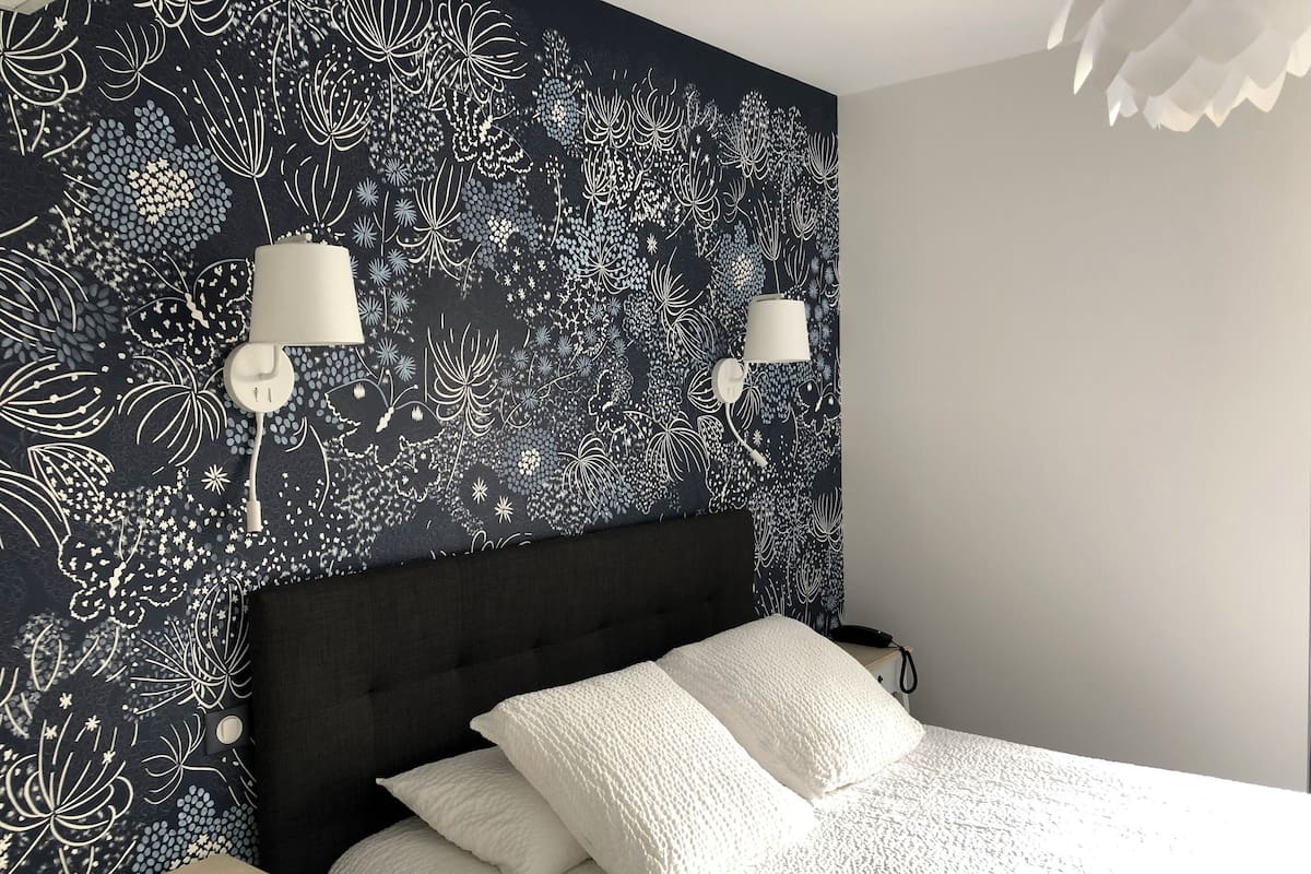Un dormitorio con papel tapiz floral oscuro, cabecero negro, ropa de cama blanca y dos lámparas de noche.