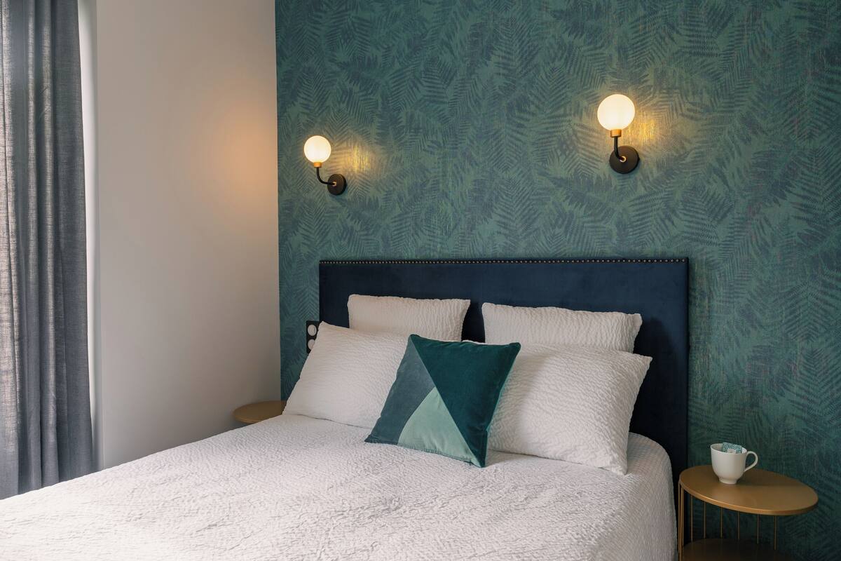 Una cama bien tendida con almohadas blancas y turquesa, cabecero oscuro y dos luces fijadas a la pared.