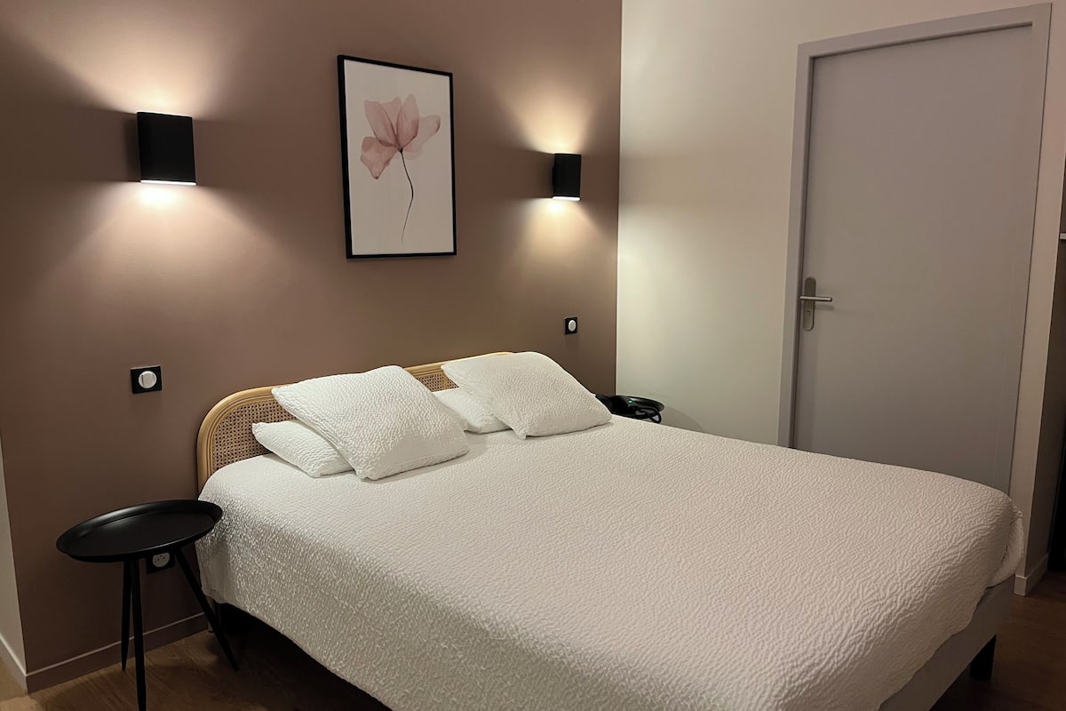 Una cama bien hecha con ropa de cama blanca, dos mesitas de noche fijadas a la pared, un cuadro enmarcado en la pared y una puerta cerrada.