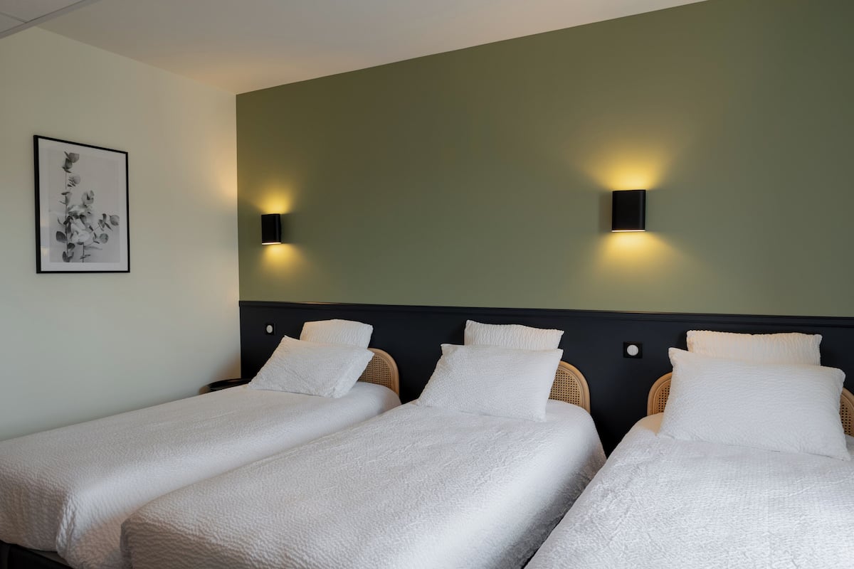 Habitación de hotel con tres camas individuales, cabecera oscura y dos luces fijadas a la pared.
