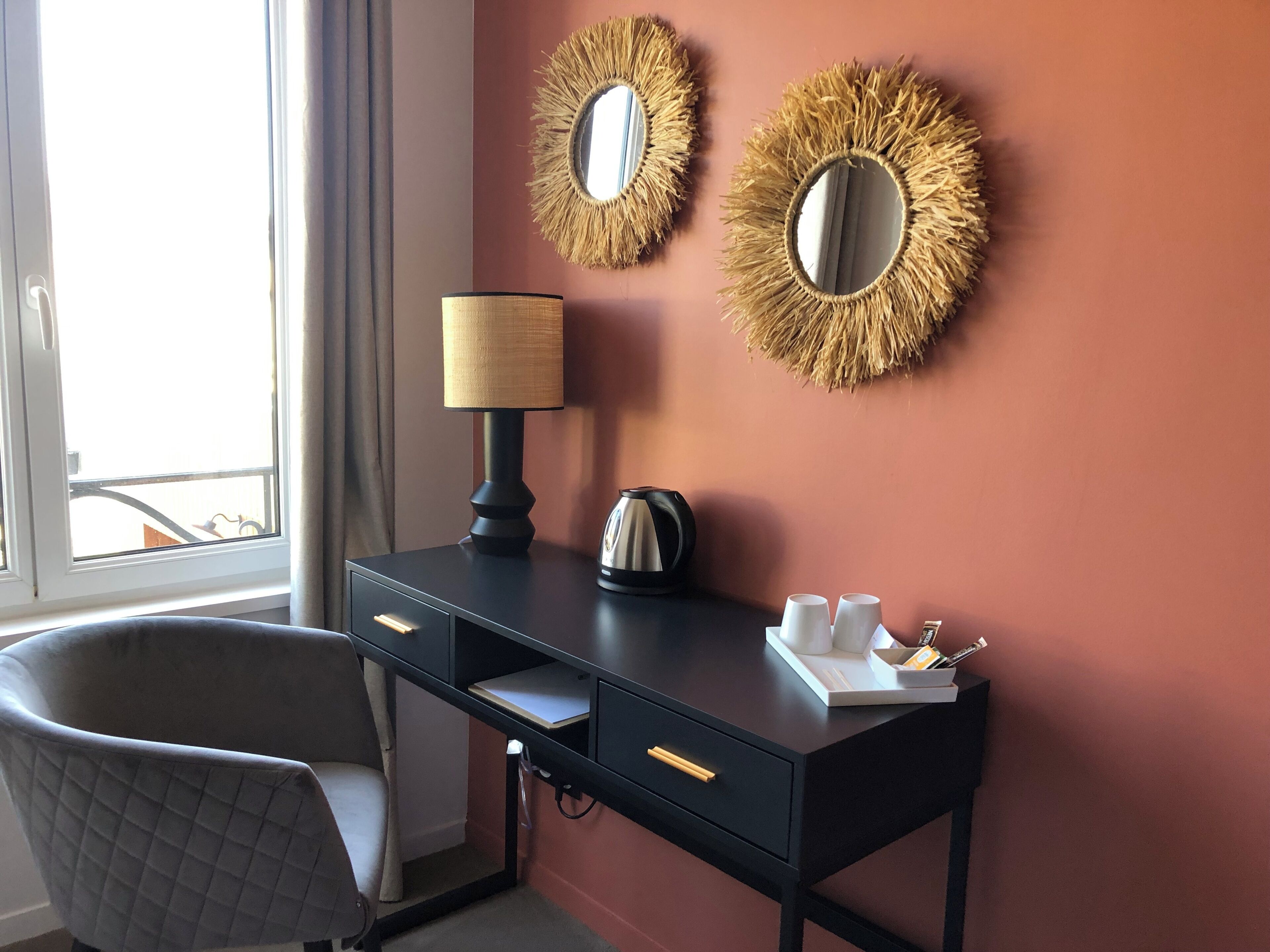 A hotel room with a desk, two wall mirrors, a lamp, a kettle, and a chair.