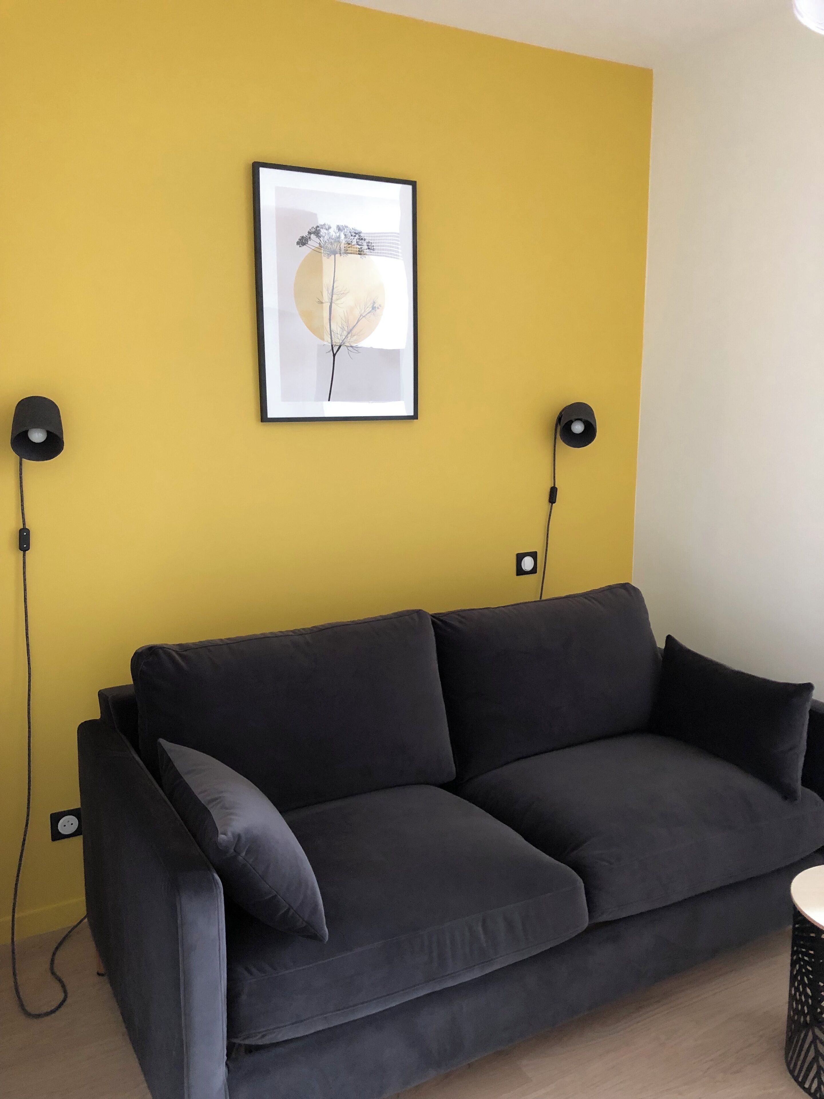 A modern living room with a dark grey sofa, two black floor lamps, and a framed picture on the yellow wall.