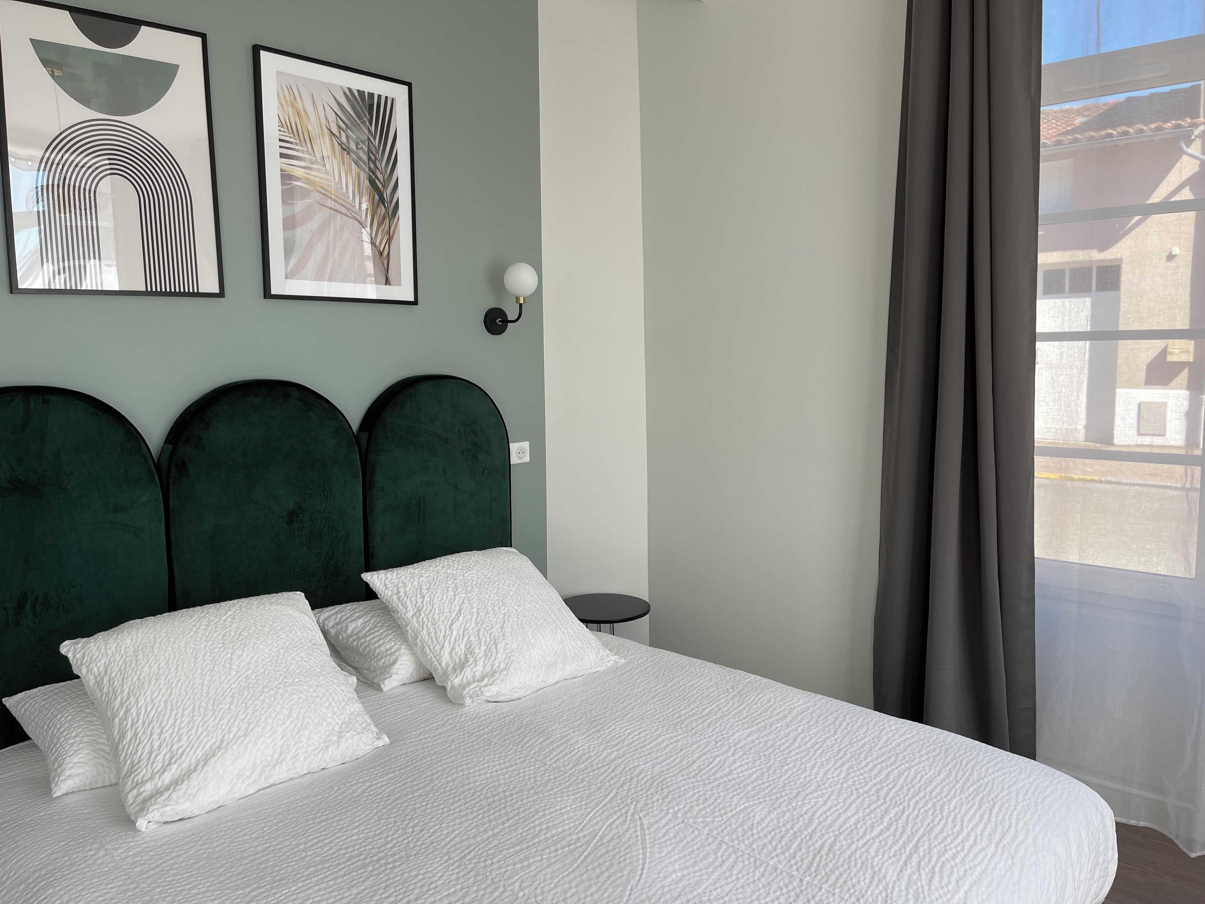 Een bed met een witte, geruite sprei en drie kussens, een donkergroen gestoffeerd hoofdbord en twee kunstwerken in lijsten aan de muur.