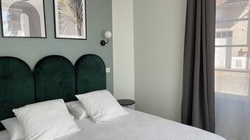Een bed met een witte, geruite sprei en drie kussens, een donkergroen gestoffeerd hoofdbord en twee kunstwerken in lijsten aan de muur.
