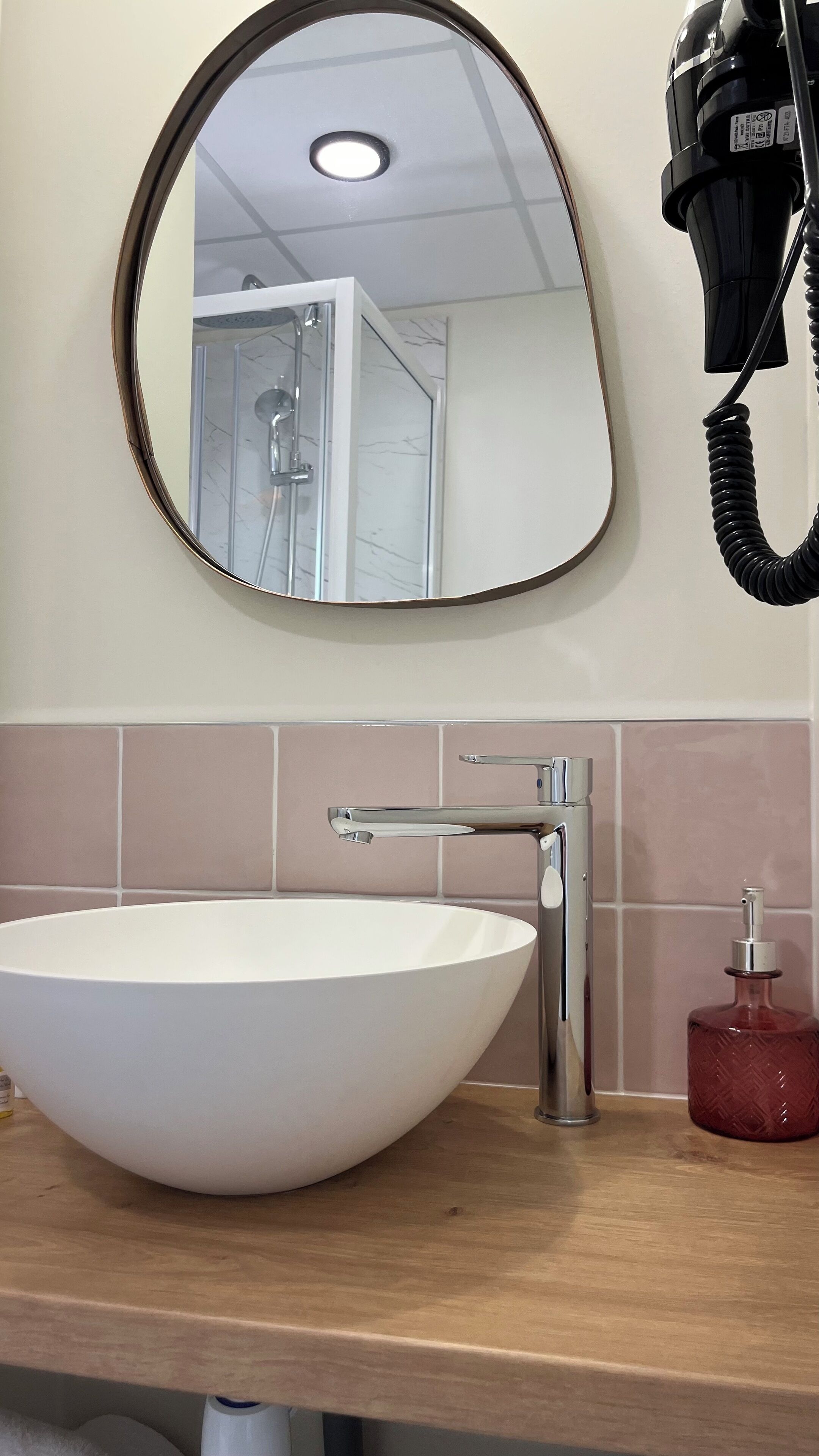 Une salle de bain avec un miroir rond, un lavabo sur colonne blanc, un sèche-cheveux et un distributeur de savon rouge.
