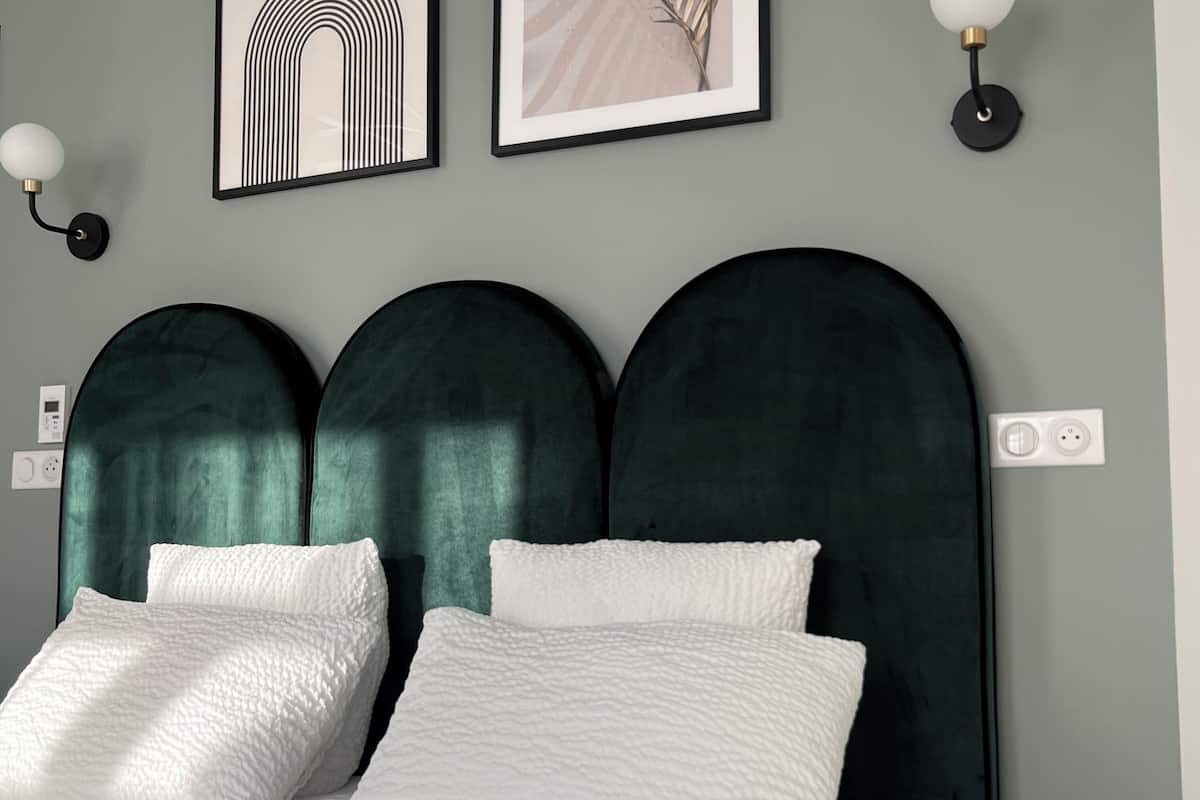 Una cama con cabecera tapizada en verde oscuro, almohadas blancas con textura y pared gris con dos cuadros enmarcados y lámparas fijadas a la pared.