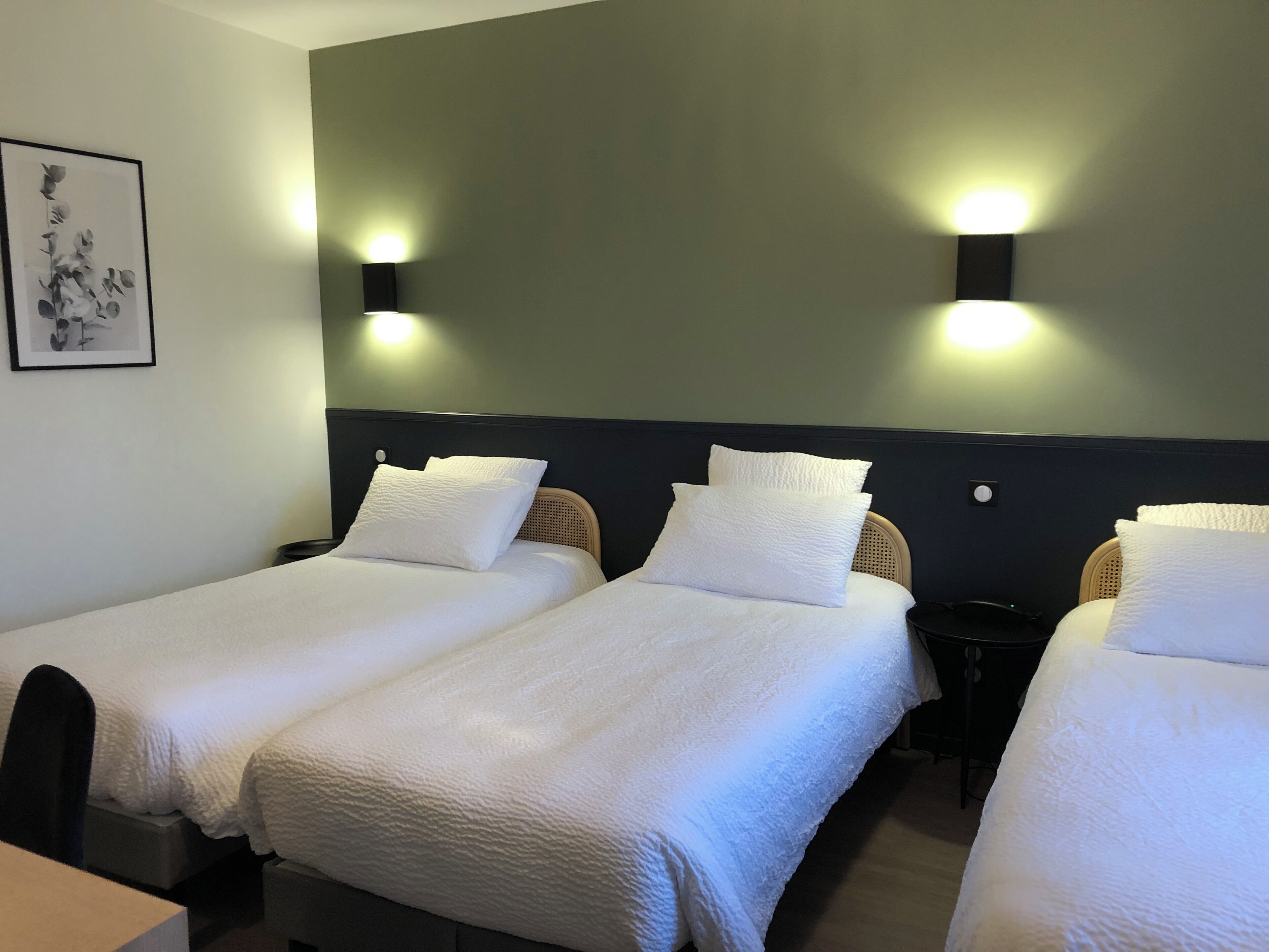Una habitación de hotel con tres camas individuales, cada una con ropa de cama blanca y cabecera. Hay luces fijadas en la pared a ambos lados de las camas.