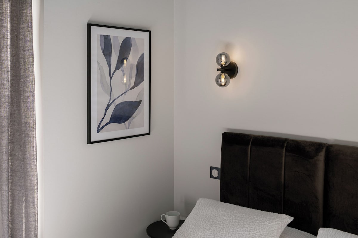 Un dormitorio con cama, una obra de arte enmarcada, una lámpara de pared y una mesita de noche con una taza.