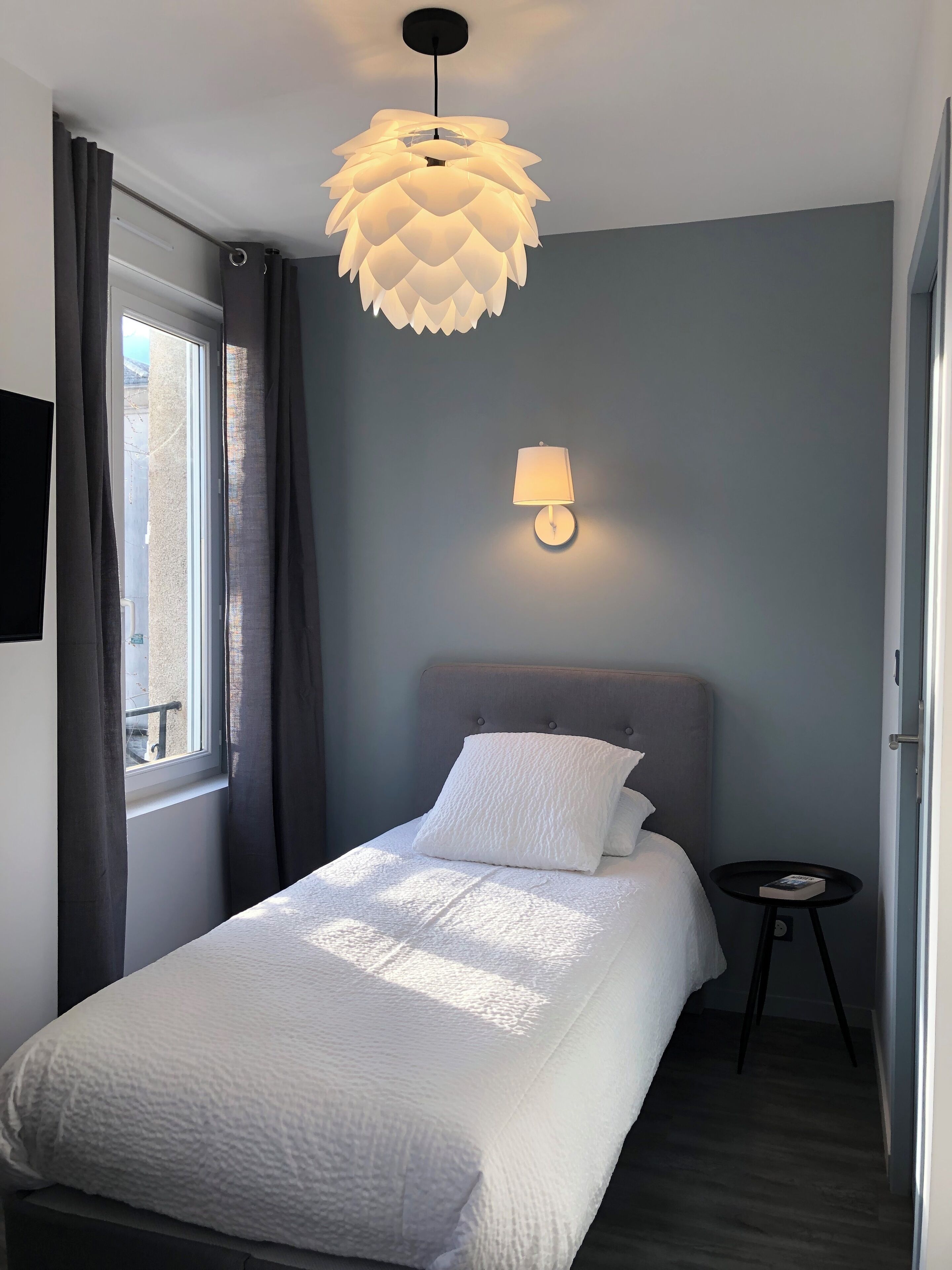 Ein ordentlich bezogenes Bett mit weißer Decke und Kissen, eine Wandlampe und eine besondere Deckenleuchte.