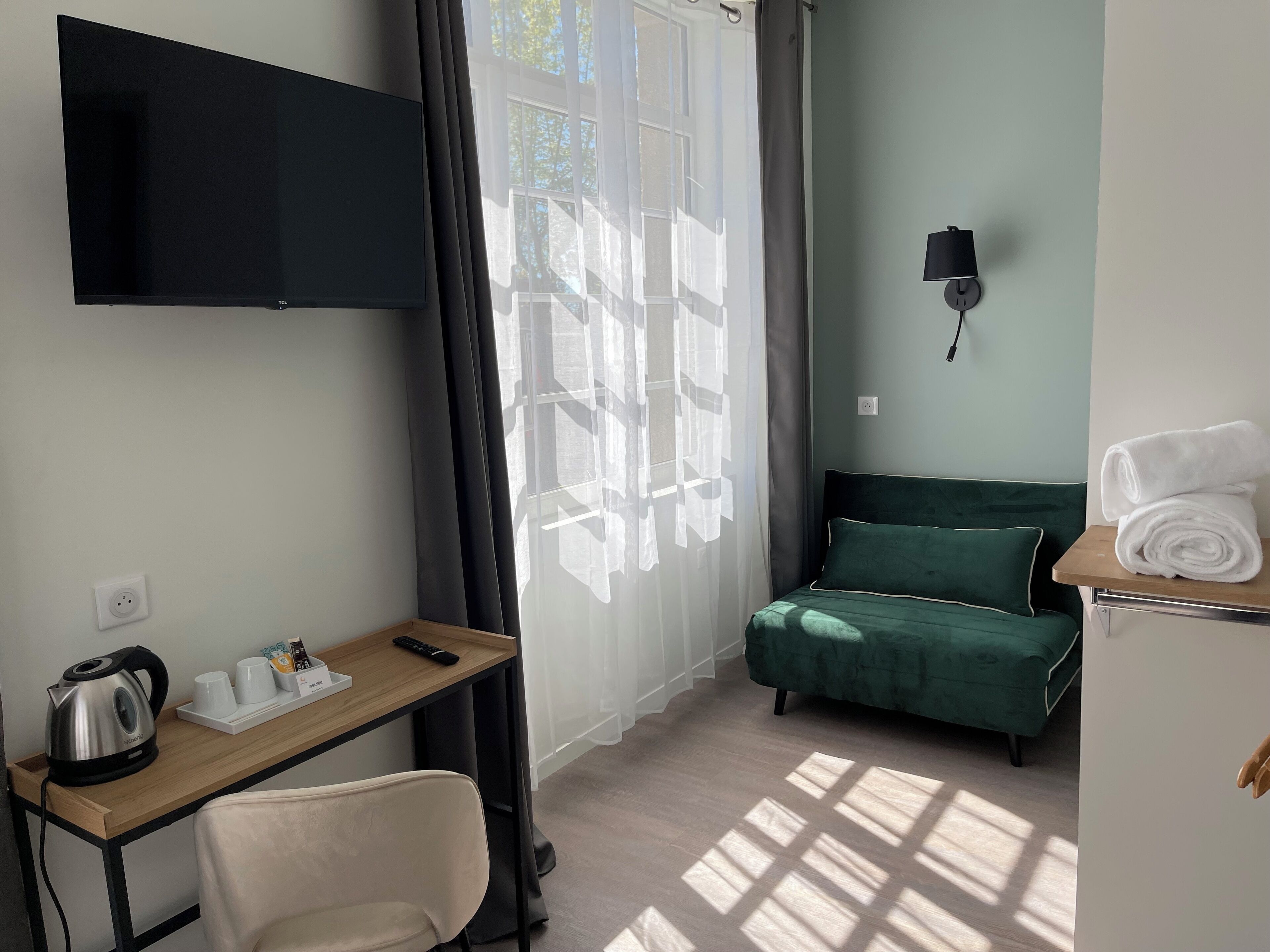 Habitación de hotel con cama, escritorio con hervidor, televisión y ventana con cortinas.