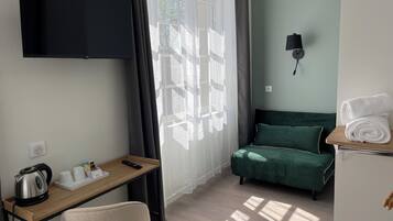 A hotel room with a bed, a desk with a kettle, a TV, and a window with curtains.