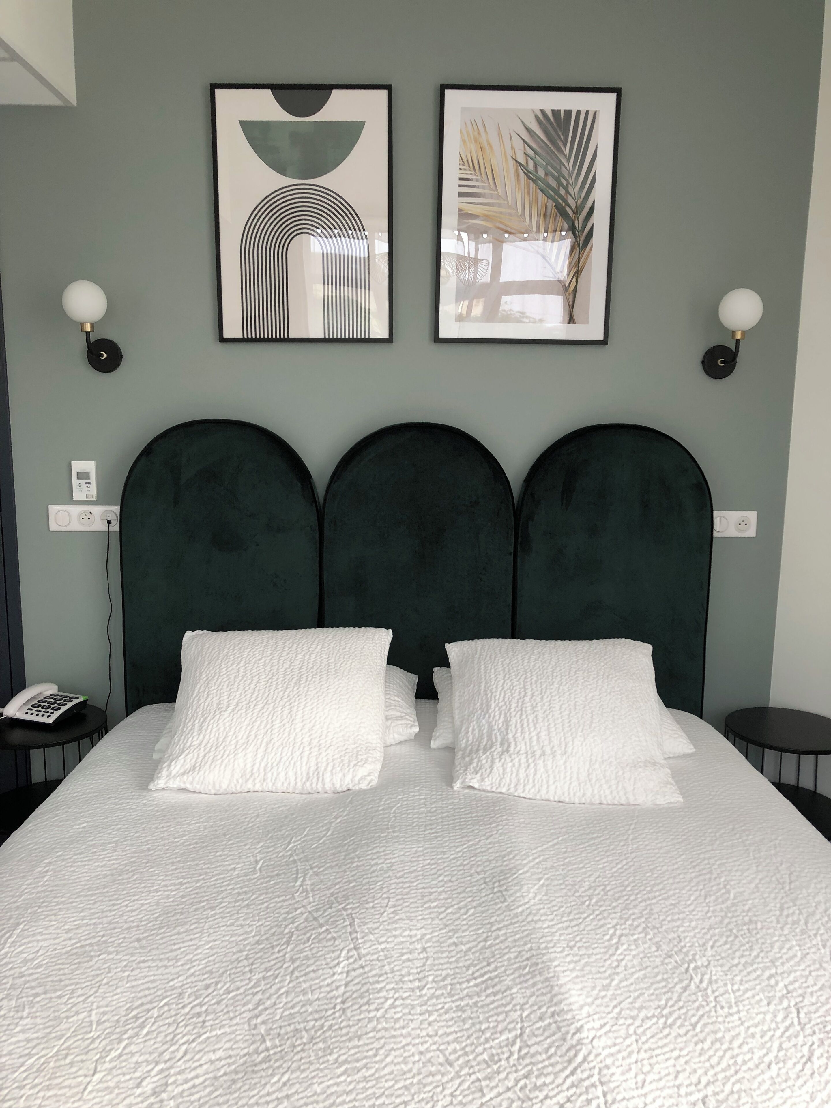 Una cama con tres cabeceros tapizados en verde oscuro, almohadillas blancas con textura y una colcha blanca acolchada. Hay dos cuadros enmarcados sobre la cama y una mesita redonda con una lámpara a la derecha.