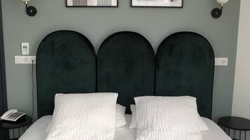 Una cama con tres cabeceros tapizados en verde oscuro, almohadillas blancas con textura y una colcha blanca acolchada. Hay dos cuadros enmarcados sobre la cama y una mesita redonda con una lámpara a la derecha.
