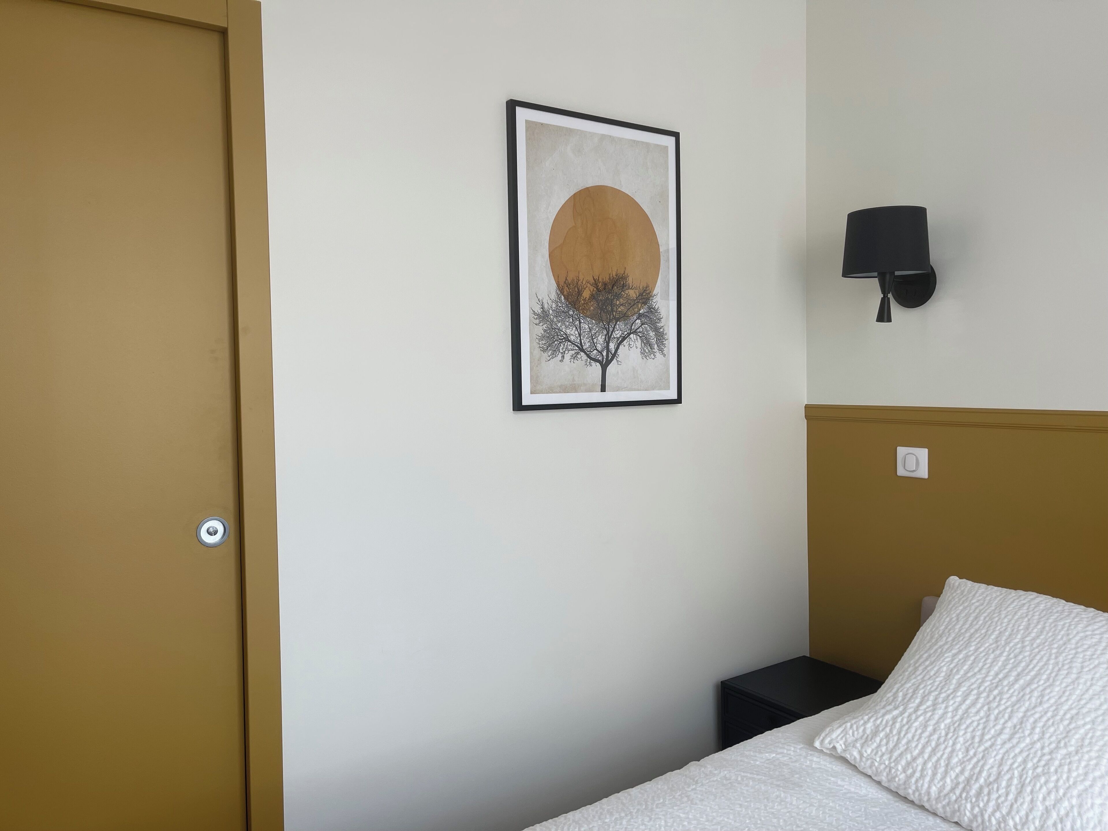 A bedroom with a bed, a wall-mounted light, a framed picture, and a door.