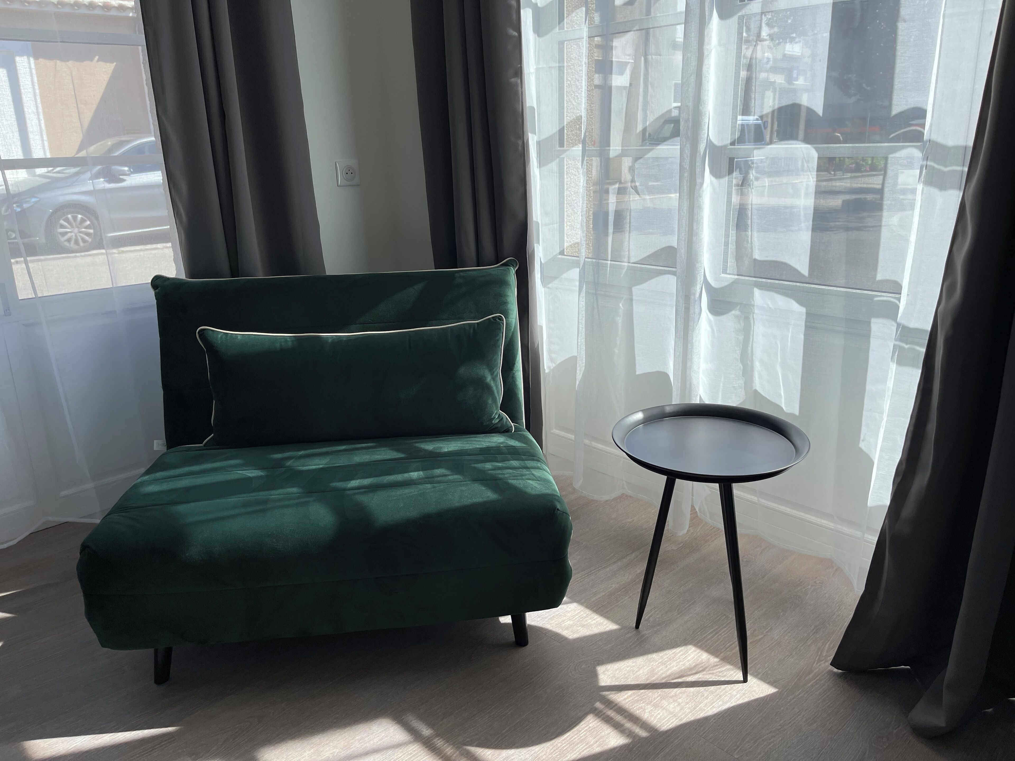 Een kamer met een groene bank, een klein rond tafeltje en uitzicht op de straat door dunne gordijnen.
