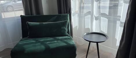 Una habitación con un sofá verde, una pequeña mesa redonda y vistas a la calle a través de cortinas traslúcidas.