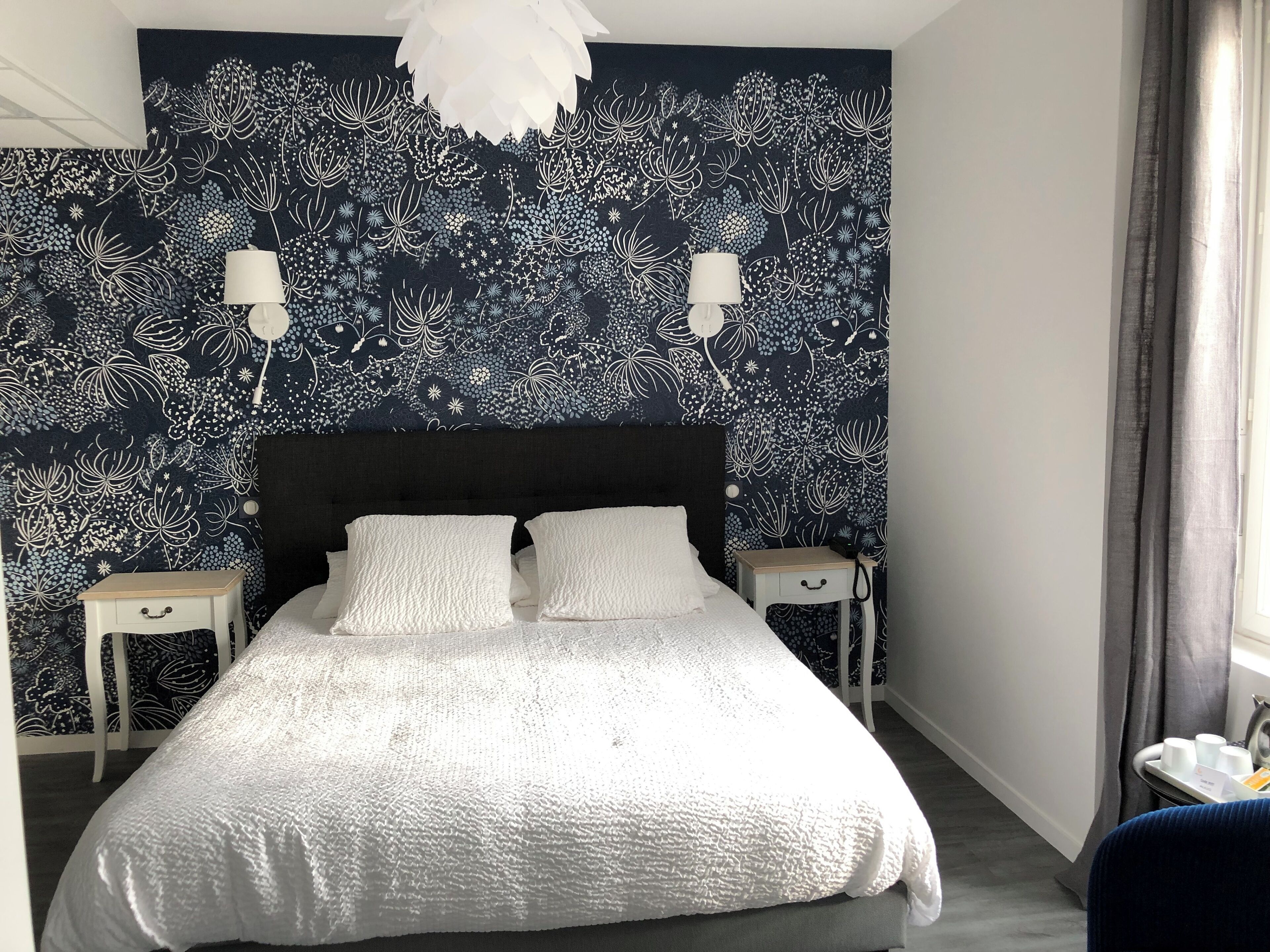 A bedroom with a dark patterned wall, a white bed, two bedside tables, and a hanging light fixture.