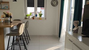 Una cocina moderna con zona de comedor, que presenta suelo de baldosas, una mesa de madera con sillas, un reloj en la pared y una ventana con cortinas.