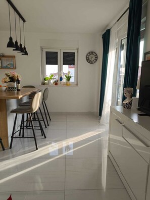 A modern kitchen with a dining area, featuring a tiled floor, a wooden dining table with chairs, a clock on the wall, and a window with curtains. - Modern apartmant 5 min walk from old town with parking (Trogir)