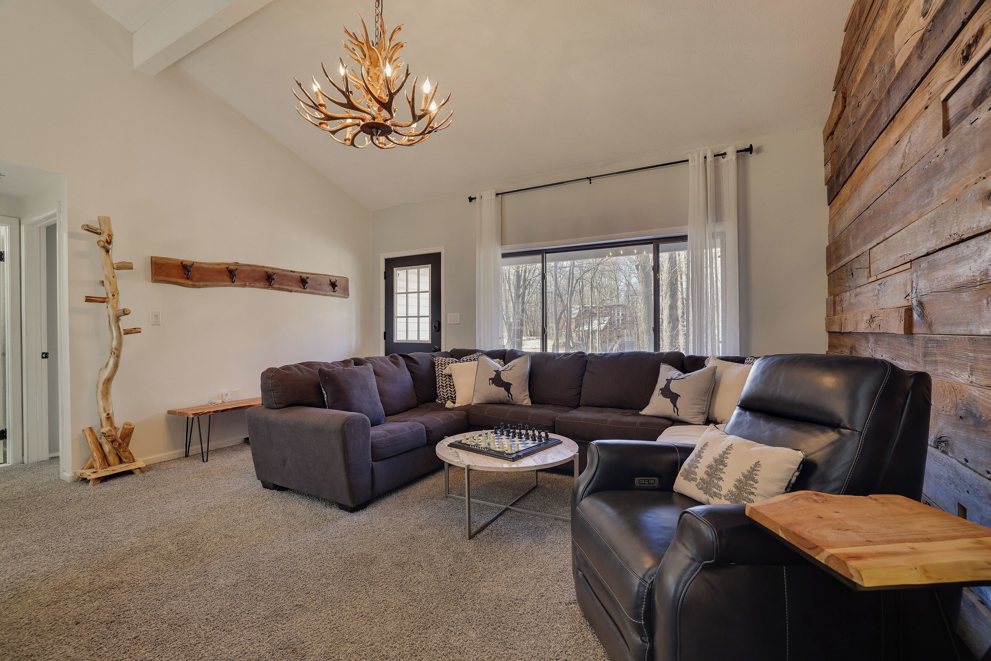 Een moderne woonkamer met een hoekbank, een leren fauteuil, een salontafel met een schaakbord, een houten wandpaneel en een kroonluchter.