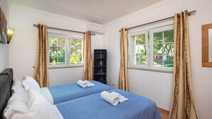 A hotel room with two beds, beige curtains, a window with a view of greenery, and an air conditioning unit.