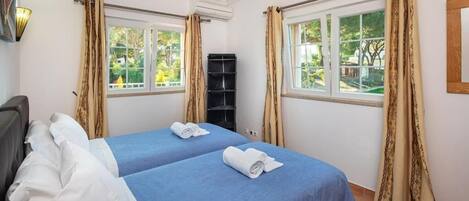 A hotel room with two beds, beige curtains, a window with a view of greenery, and an air conditioning unit.