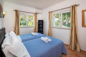 Habitación de hotel con dos camas, cortinas color beige, una ventana con vista a follaje y un equipo de aire acondicionado.