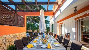 An outdoor dining area with a long table set for a meal, surrounded by wicker chairs, under a pergola with a white umbrella.