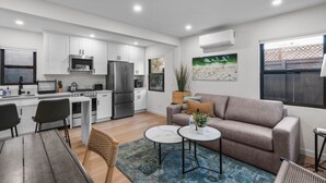 A modern kitchen with white cabinets, stainless steel appliances, and a dining area with a wooden table and chairs. - Pacific Beach Surf Nook Blocks to Surf & Sand (San Diego)