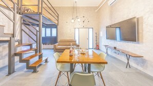 A modern living space with a wooden dining table, light-colored chairs, a sofa, and a staircase with metal railings.